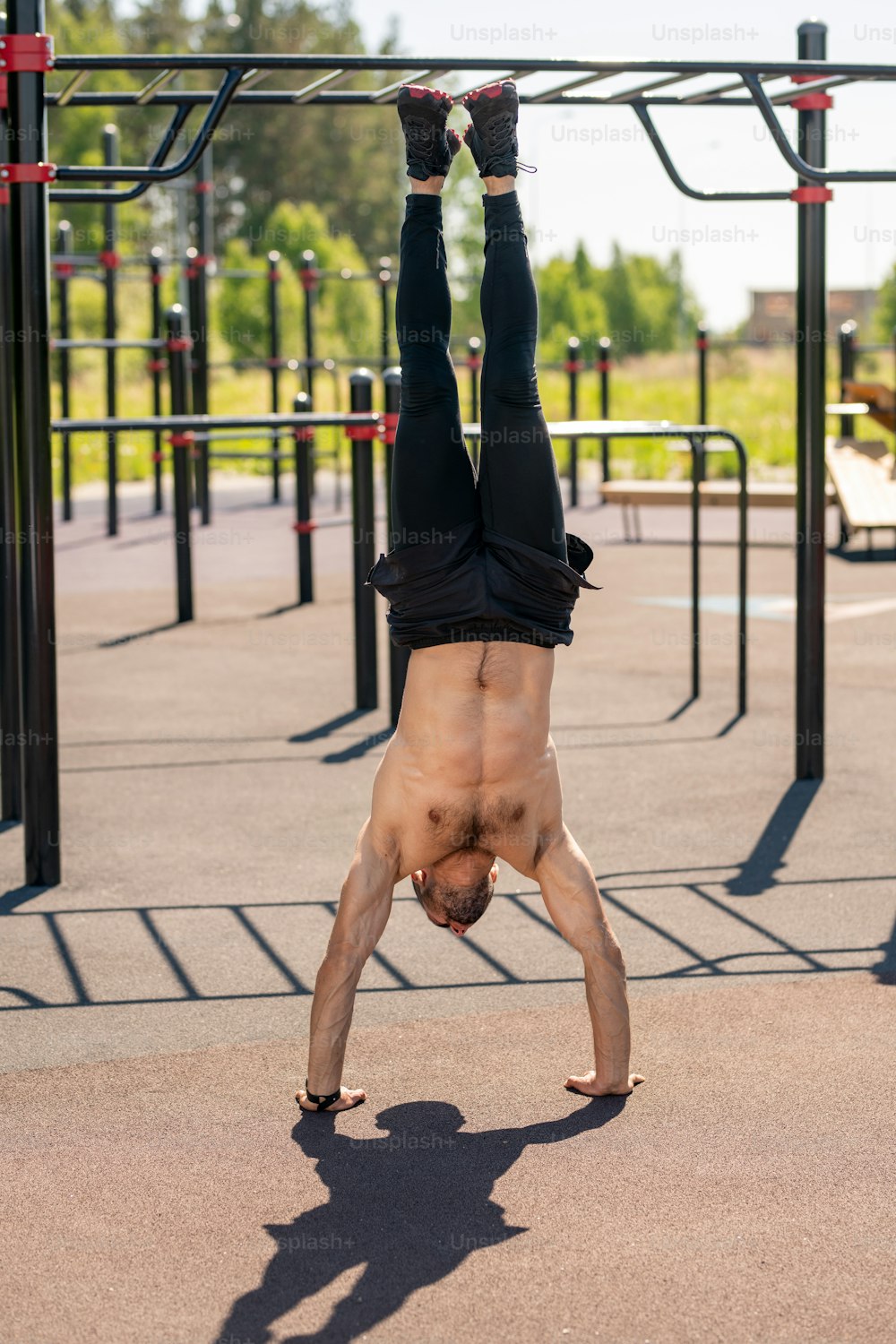 Front view of young shirtless muscular athlete doing handstand while exercising on sportsground