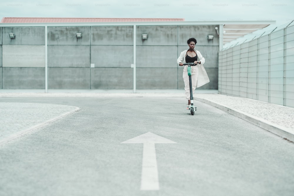 Uma jovem mulher africana de óculos e um manto branco esvoaçante está andando de patinete elétrico na estrada de asfalto com uma seta pintada como uma marcação de estrada em um primeiro plano desfocado, profundidade de campo rasa