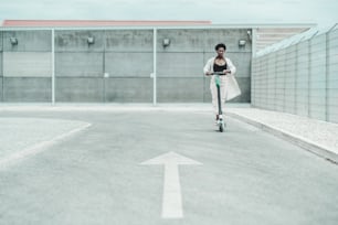 Eine junge afrikanische Fancy Frau mit Brille und weißem flatterndem Umhang fährt auf einem E-Scooter auf der Asphaltstraße mit einem aufgemalten Pfeil als Straßenmarkierung im unscharfen Vordergrund, geringe Tiefenschärfe