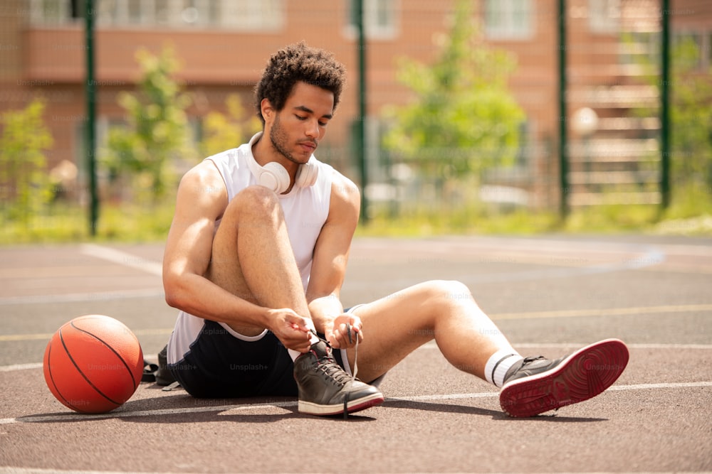 Junger gemischtrassiger Athlet in Sportkleidung sitzt auf dem Basketballplatz und bindet vor dem Training einen Schnürsenkel aus Turnschuhen