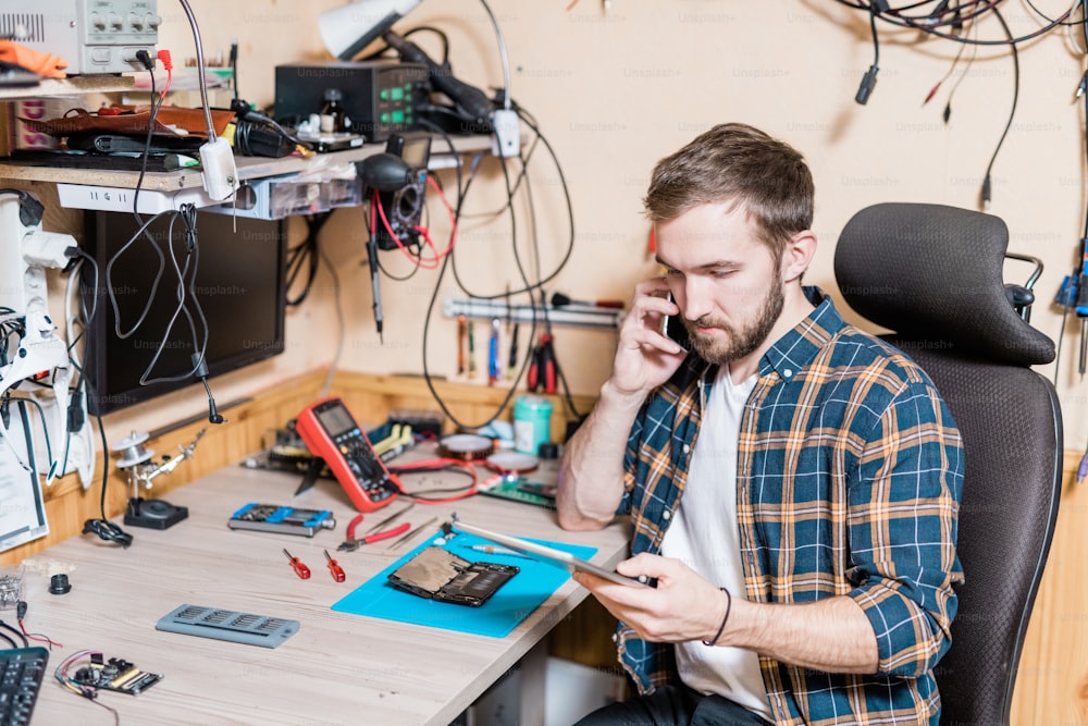 Junger professioneller Meister des Gadget-Reparaturdienstes, der mit dem Kunden auf dem Smartphone spricht, während er Online-Anfragen im Touchpad durchsucht