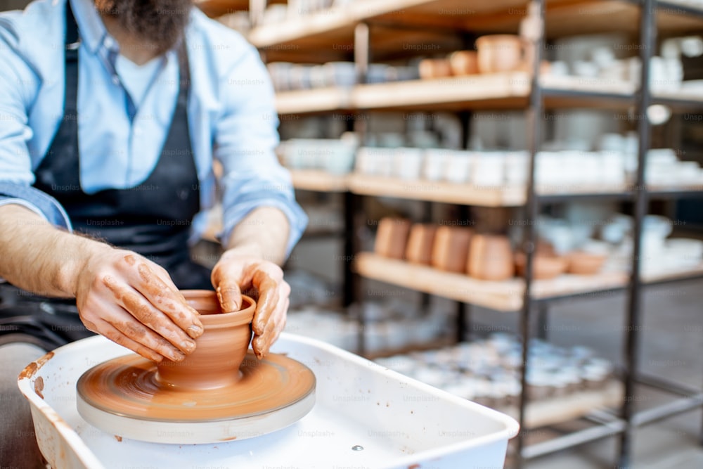 Homem que faz jarro de barro formando forma pelas mãos na roda de cerâmica dentro de casa, vista de close-up