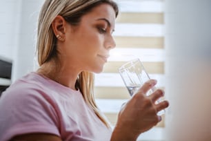 Profile of beautiful caucasian blond woman standing next to window in kitchen and drinking water.