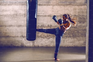 Joven en forma atractiva musculosa caucásica kick boxer en ropa deportiva con guantes de boxeo pateando bolsa de boxeo.