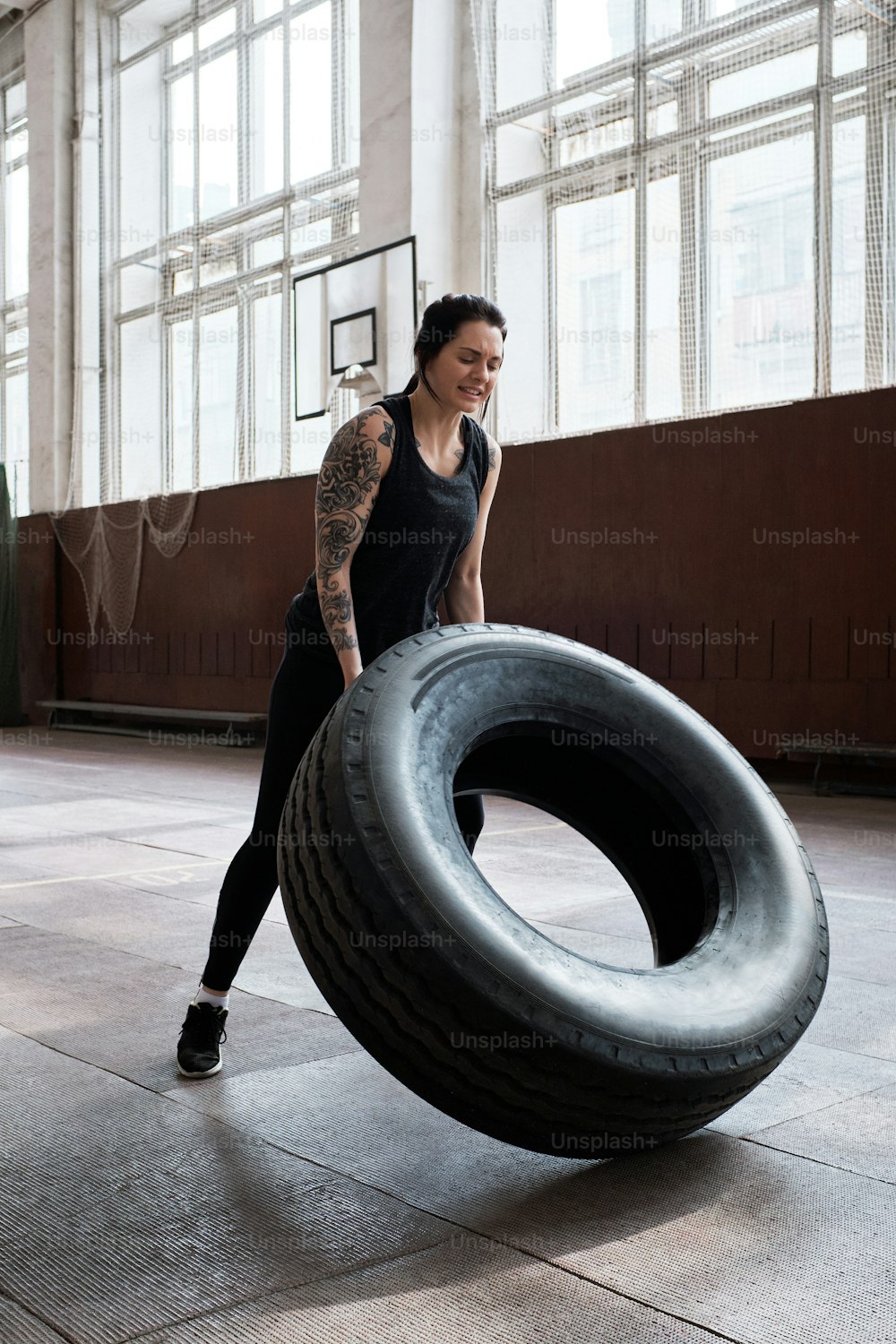 Athletic tattooed Caucasian woman in black sportswear flipping heavy tire during cross training in sports hall