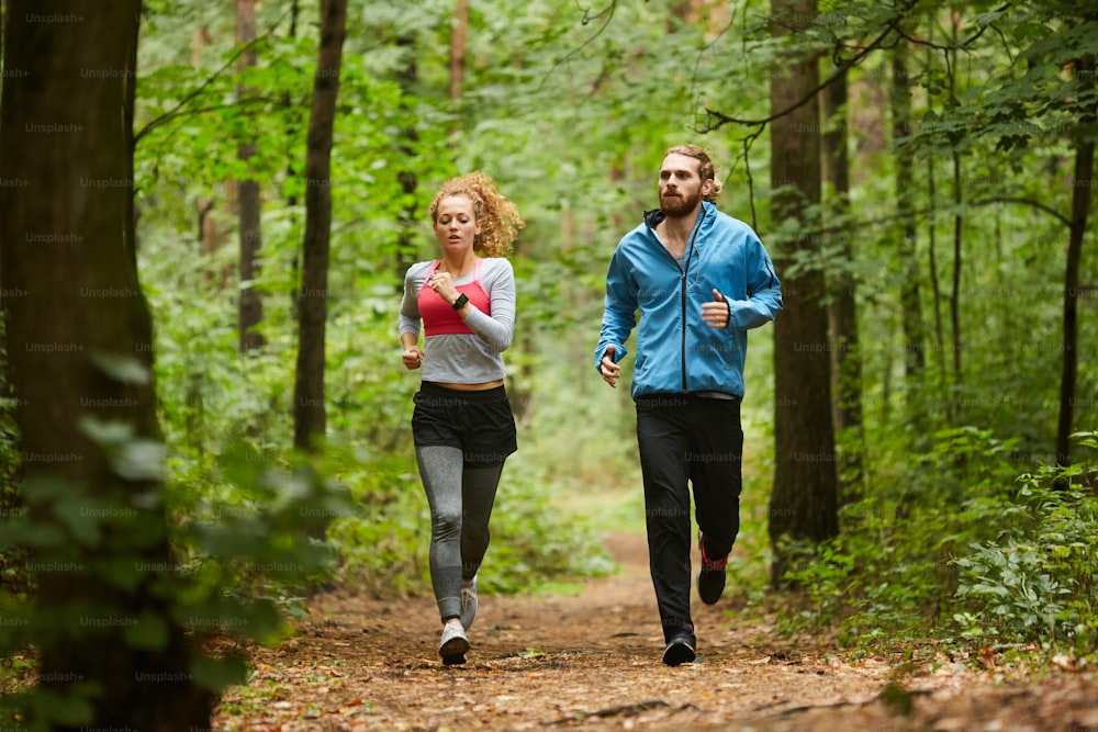 Giovane coppia sportiva che corre lungo il sentiero forestale o nel parco al mattino tra alberi verdi