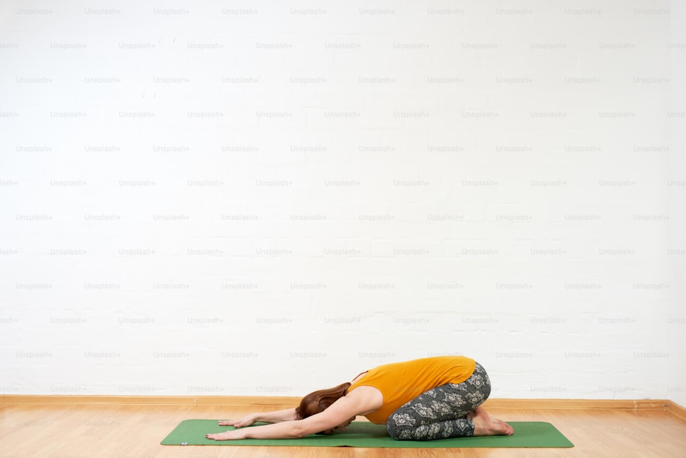 Full length image of middle age woman doing yoga flow on green mat near white wall, she is wearing yellow tank and dark pants