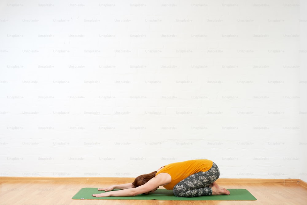 Imagem de corpo inteiro da mulher de meia-idade que faz o fluxo de yoga no tapete verde perto da parede branca, ela está vestindo tanque amarelo e calças escuras