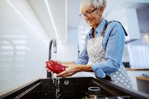 Atraente mulher loira sênior caucasiana feliz no avental em pé na cozinha e lavando pimenta vermelha na pia da cozinha.