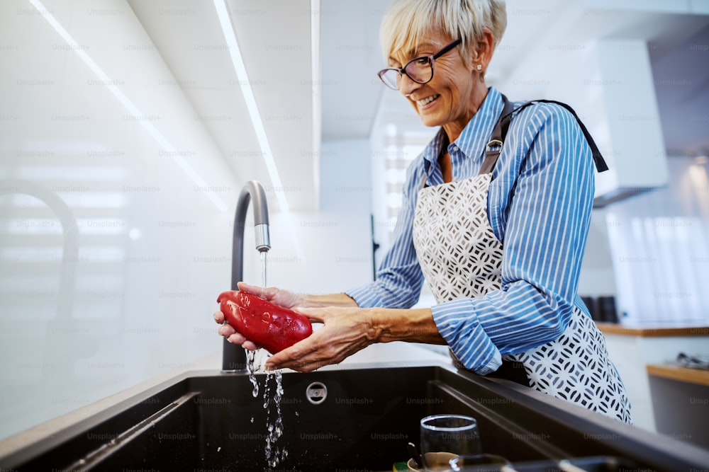 Attraktive glückliche kaukasische blonde Frau in Schürze, die in der Küche steht und rote Paprika in der Küchenspüle wäscht.