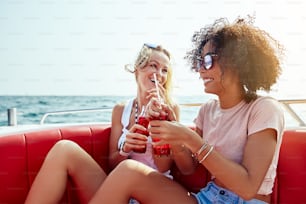 Two young female friends sitting on a boat on the open ocean sipping on drinks together during their summer vacation