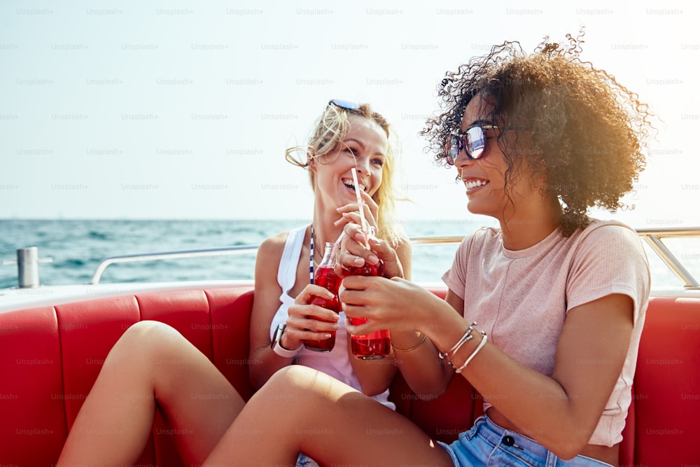 Dos jóvenes amigas sentadas en un bote en el océano abierto bebiendo bebidas juntas durante sus vacaciones de verano