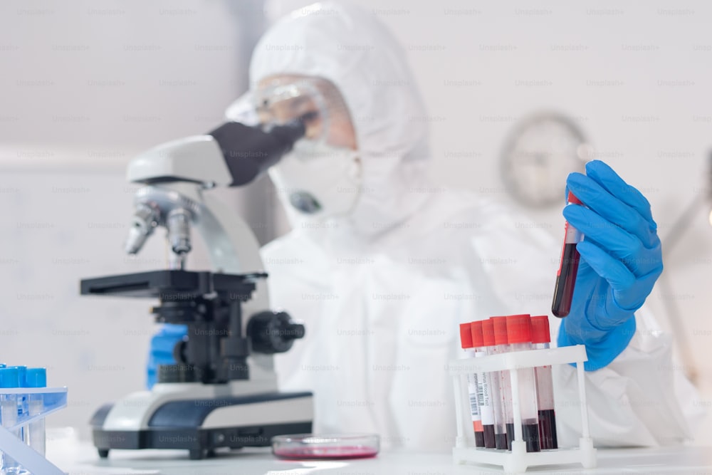 Close-up of lab technician in protective suit examining blood samples under microscope in laboratory