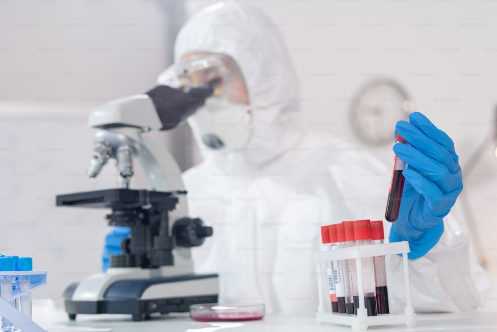 Close-up of lab technician in protective suit examining blood samples under microscope in laboratory