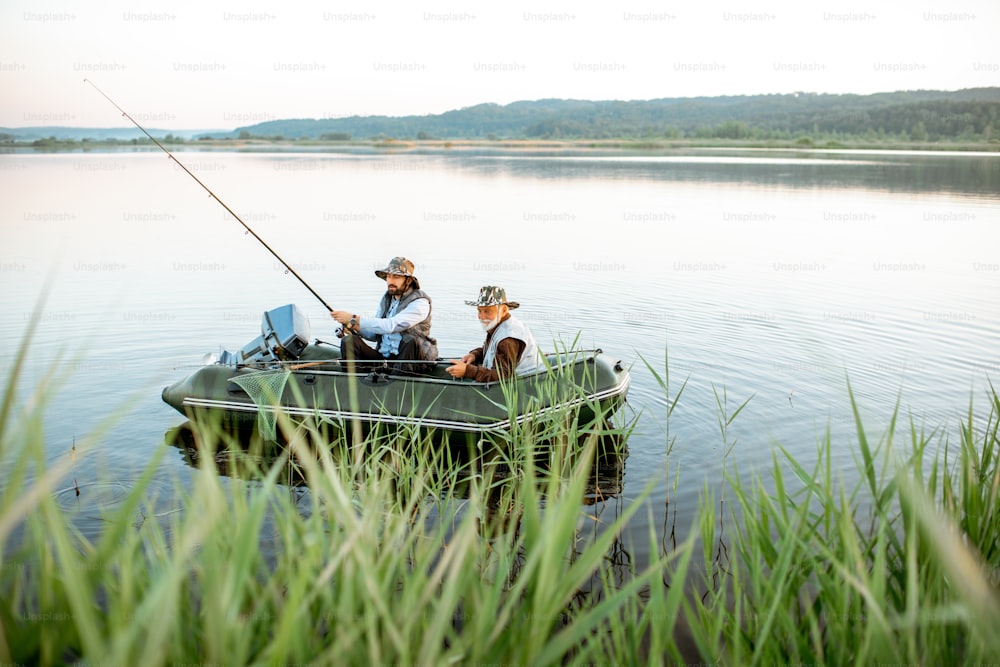 Grandfather with adult son fishing on the inflatable boat on the lake with green cane on the foreground early in the morning