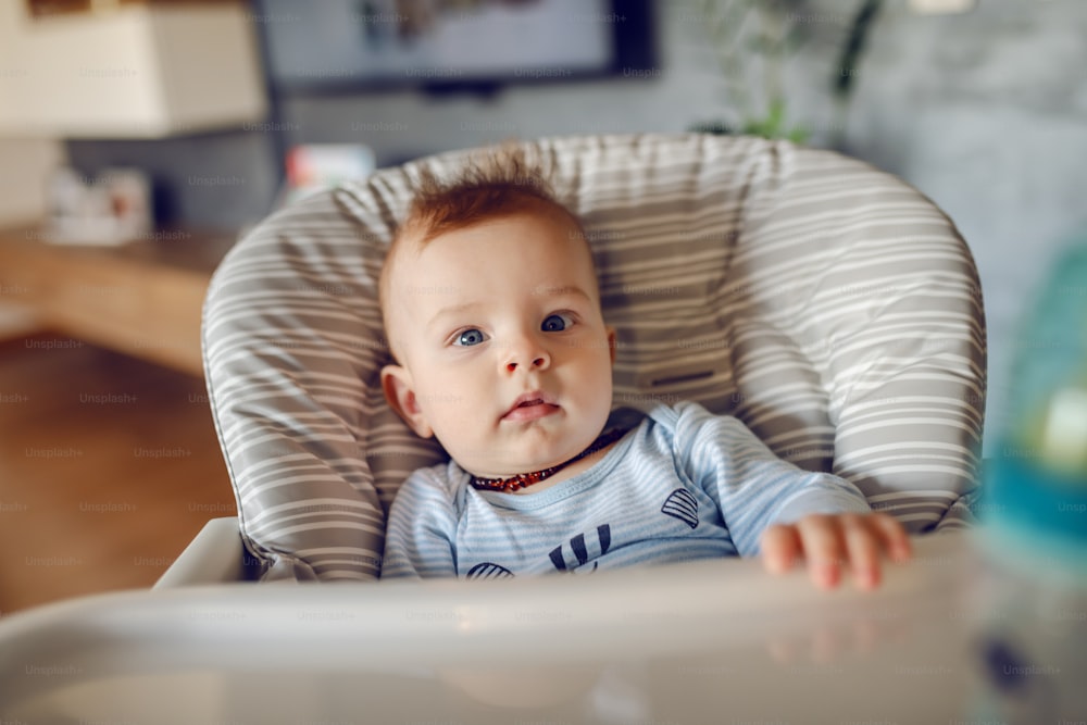 Beautiful caucasian curious baby boy sitting in his chair at home and waiting to be feed.