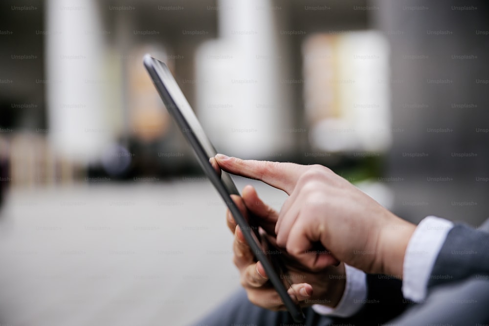 Closeup of man in suit and scrolling on tablet outdoors. Selective focus on hand.