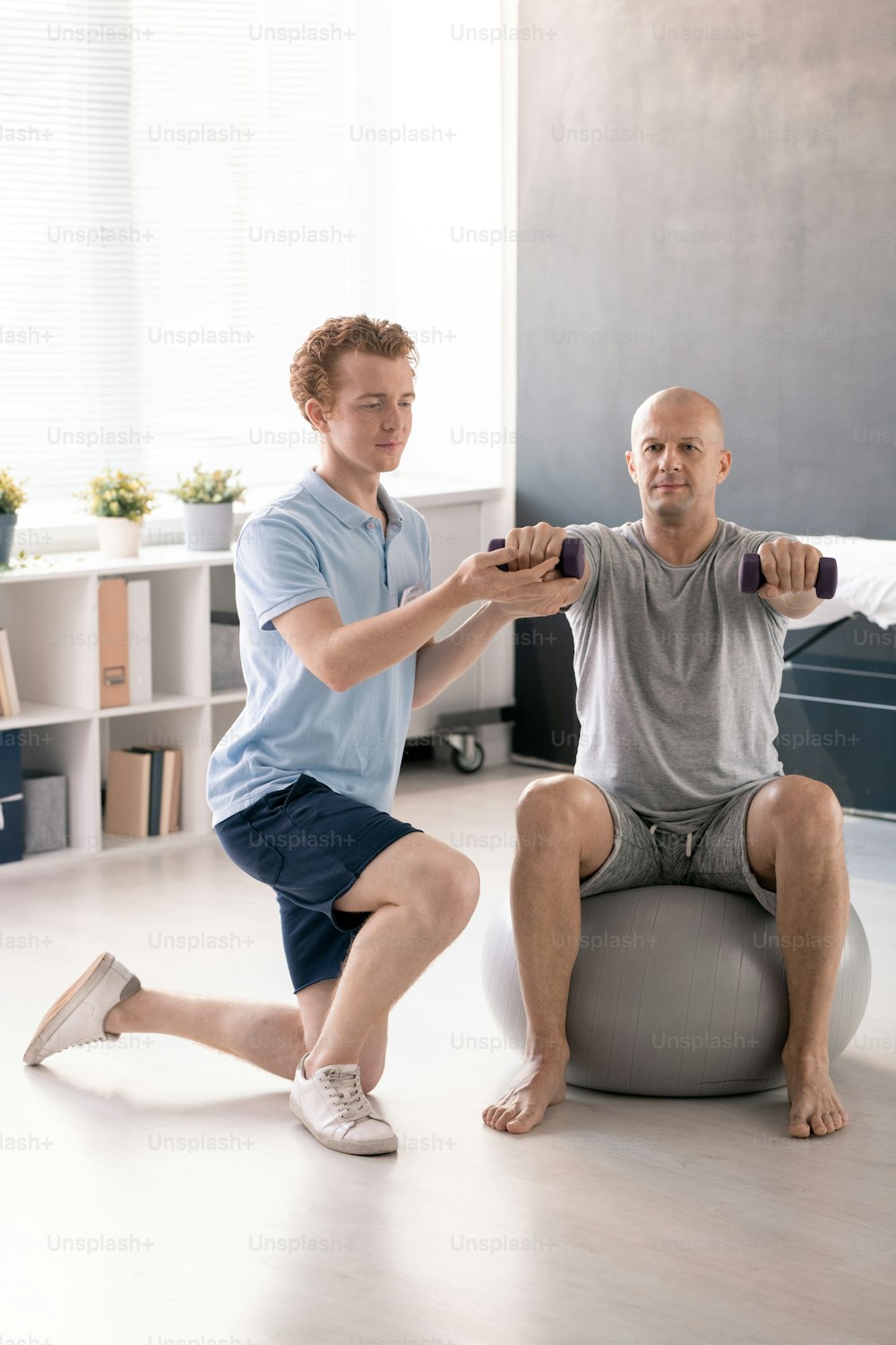 Joven fisioterapeuta ayudando a un paciente masculino a hacer ejercicio con mancuernas y apoyando su brazo derecho durante el entrenamiento en fitball en clínicas