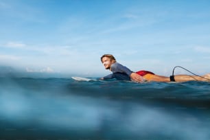 Surfe. Homem surfista no retrato da prancha de surfe. Cara bonito em traje de mergulho nadando no oceano. Mar turquesa e céu azul no fundo.
