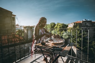 An adorable young black woman is having lunch and cutting into pieces delicious pizza with cheese and bacon while sitting on a balcony of a cozy outdoor restaurant with a glass of wine on the table