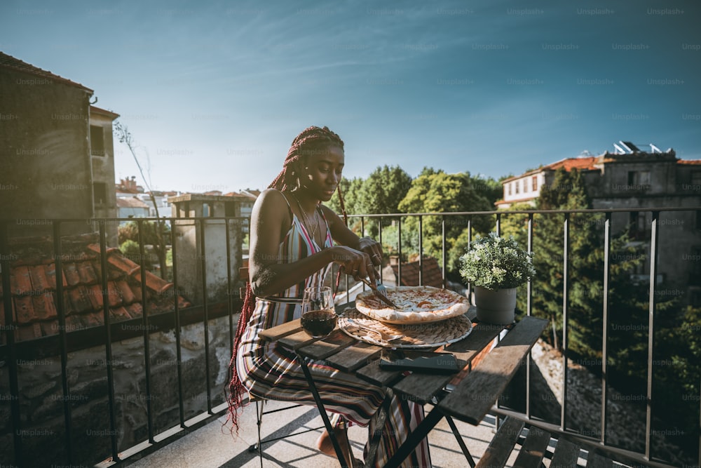 An adorable young black woman is having lunch and cutting into pieces delicious pizza with cheese and bacon while sitting on a balcony of a cozy outdoor restaurant with a glass of wine on the table
