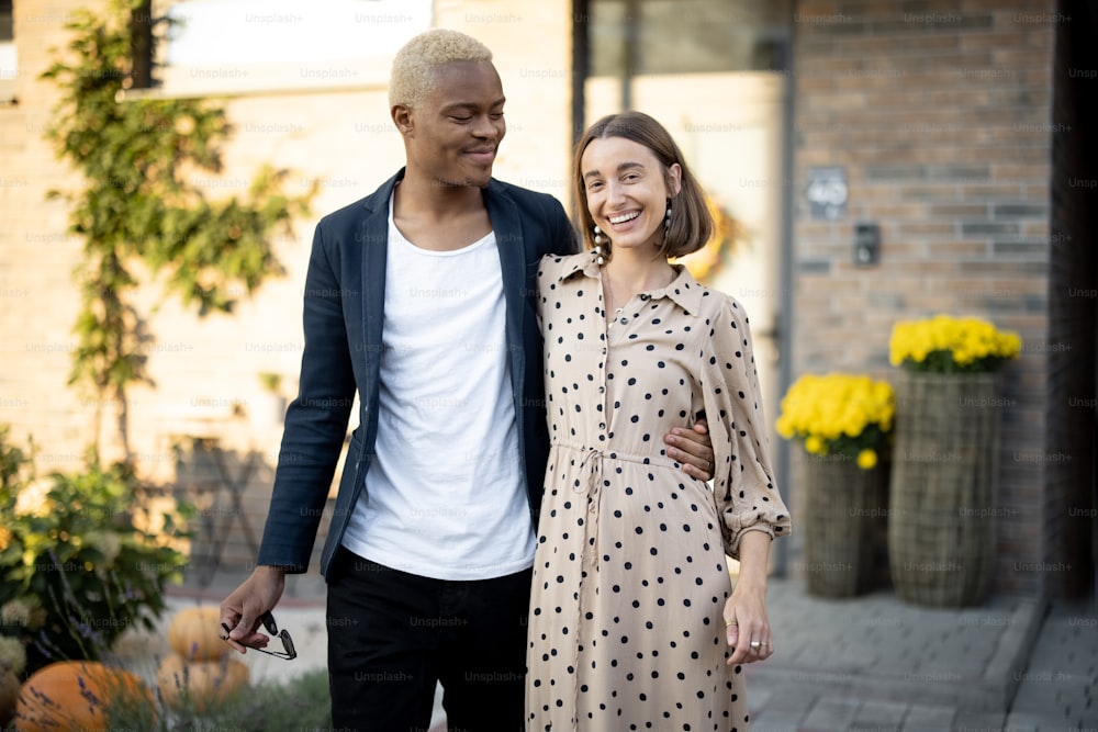 Multiracial couple going together on background of their house. Concept of relationship and enjoying time together. Young black man embracing his caucasian girlfriend. Modern lifestyle. Sunny daytime