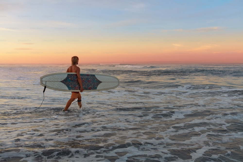 Wellenreiter. Surfer Mann mit weißem Surfbrett, das im Ozean spazieren geht. Wassersport für einen aktiven Lebensstil. Wunderschöner tropischer Sonnenuntergang auf See.