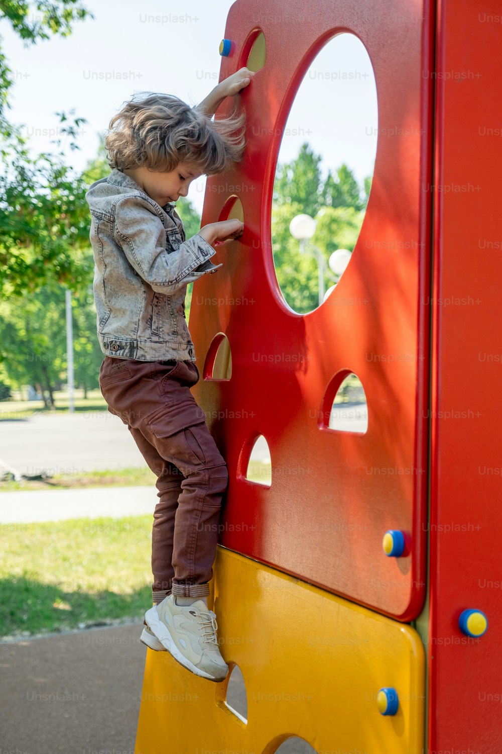 Garotinho bonito em sapatos cruzados, jaqueta jeans e calças subindo ou descendo em instalações de lazer para crianças no parque no dia de verão