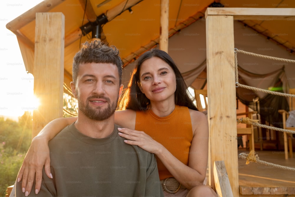 Happy young brunette woman embracing her husband while both enjoying summer rest by glamping house