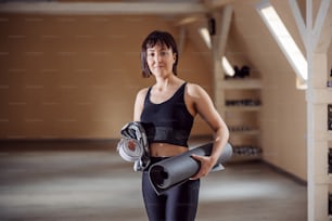 Fit slim smiling sportswoman standing in fitness studio, holding mat and fitness equipment.