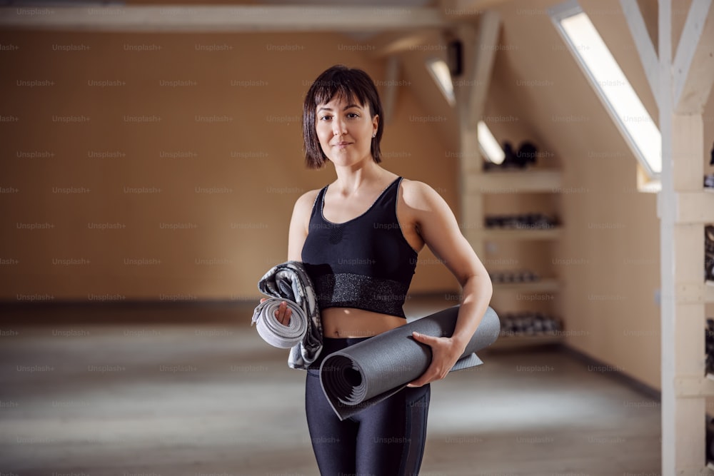 Fit slim smiling sportswoman standing in fitness studio, holding mat and fitness equipment.