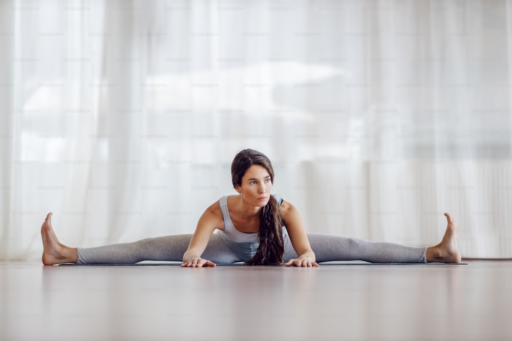 Young attractive brunette sitting on the mat with spread legs and bending forward. Healthy life concept.