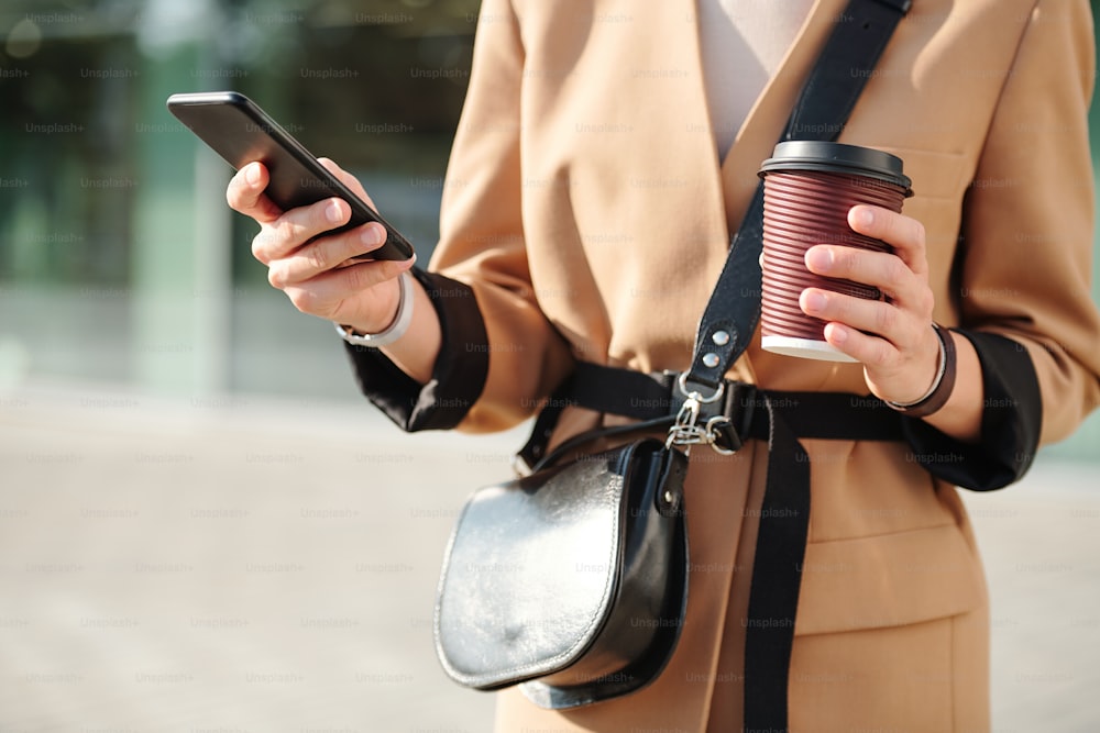Manos de una joven mujer elegante con bebida y bolso desplazándose por los contactos en el teléfono inteligente mientras va a llamar a alguien al aire libre
