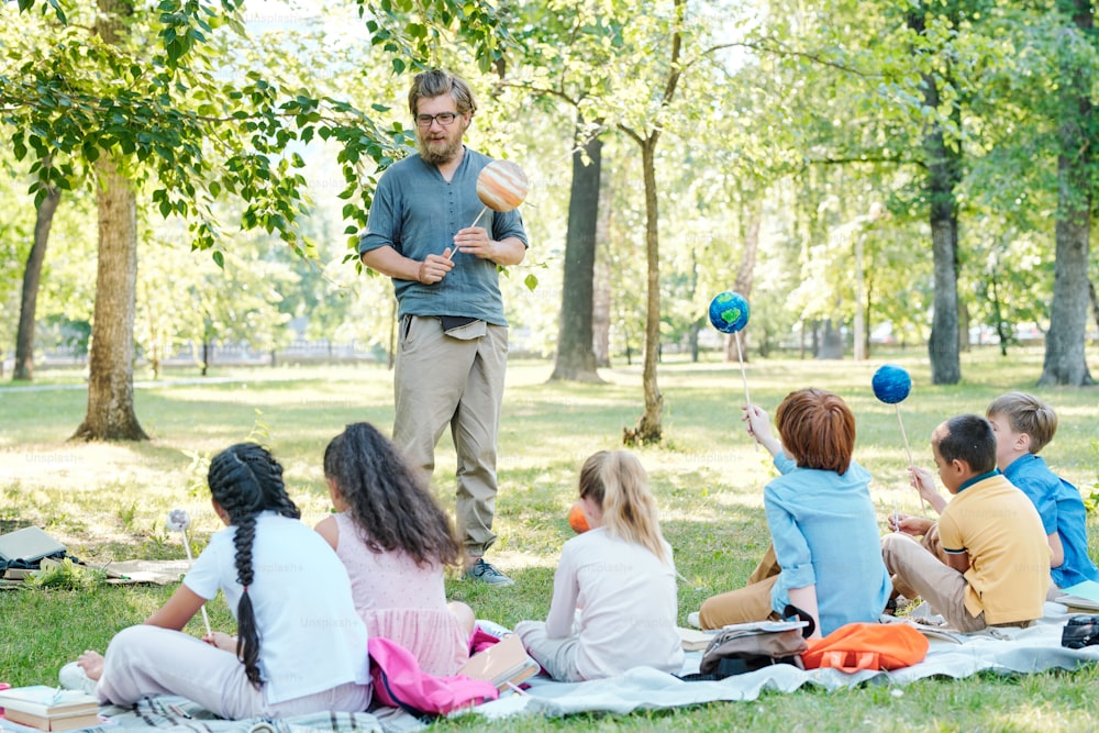 Bearded teacher holding planet model on stick and talking to group of children sitting on ground in park
