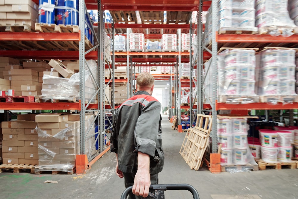 Vista trasera del joven trabajador rubio del almacén en uniforme tirando del carro con mercancías mientras se mueve hacia el pasillo entre estanterías con paquetes