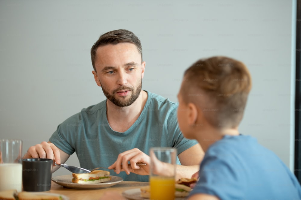 Serious young man eating homemade sandwich for breakfast and looking at his little son while discussing plans for the day with him