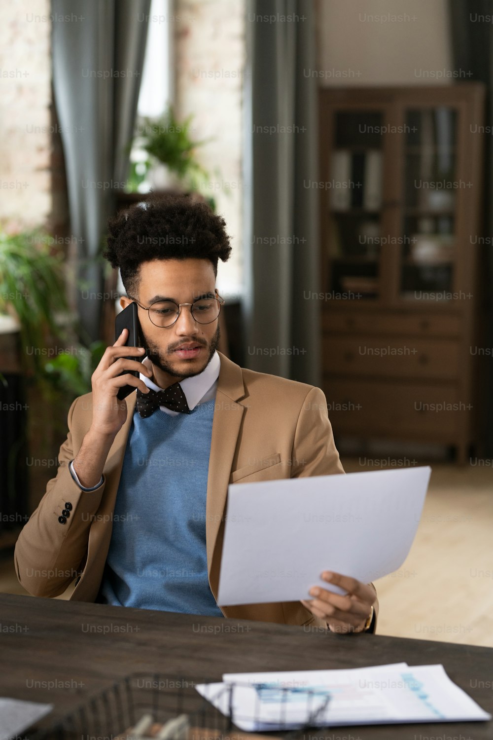 Young man sitting at his workplace talking on mobile phone and examining business contract at office