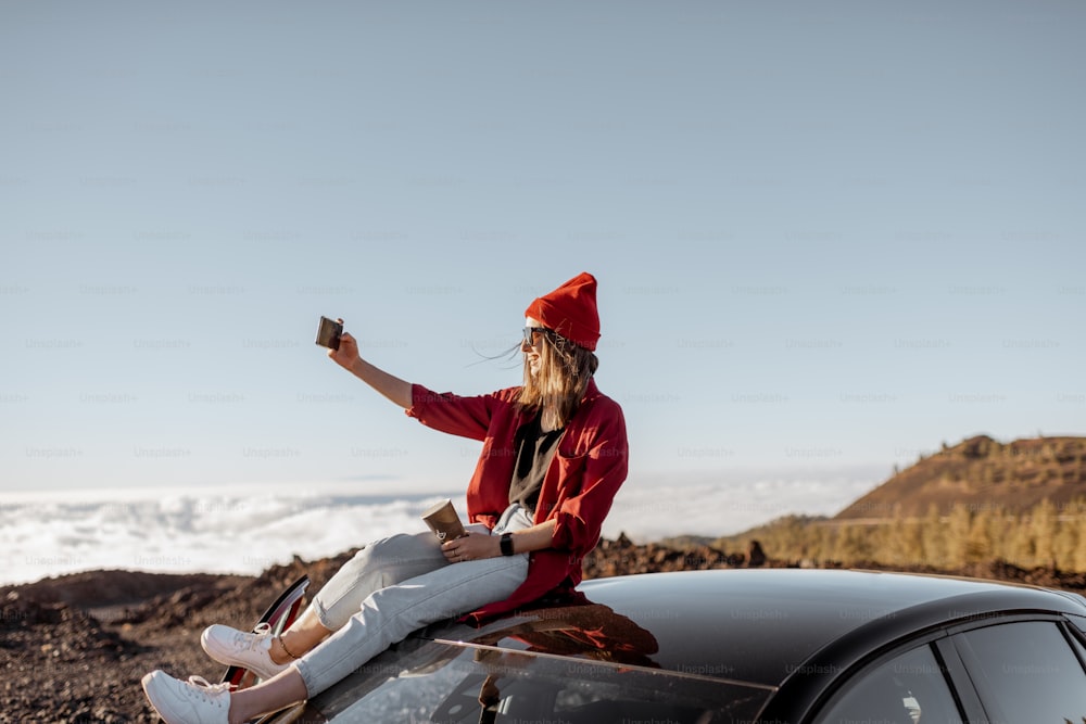 Jovem mulher vestida de vermelho desfrutando de paisagens rochosas acima das nuvens, de pé no carro altamente nas montanhas em um pôr do sol. Estilo de vida despreocupado e conceito de viagem