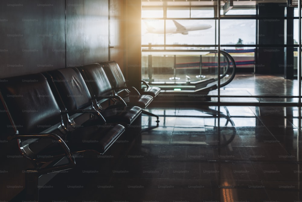 Uma sala de espera vazia de uma moderna área de embarque ou desembarque do terminal do aeroporto com uma fileira de poltronas de metal de couro e um viajante à distância; um avião decolando do lado de fora da janela