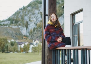 Young woman in coat sitting on wooden fence near cottage on mountains background