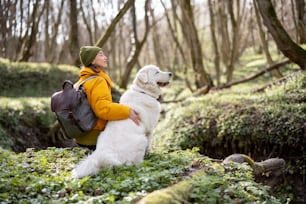 Young woman in hiking clothes and backpack sitting near big white dog in green spring forest. Enjoys and explore of tranquil nature. Look aside.