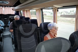 Aged Caucasian woman with grey hair sitting by window inside bus and looking at road and country houses