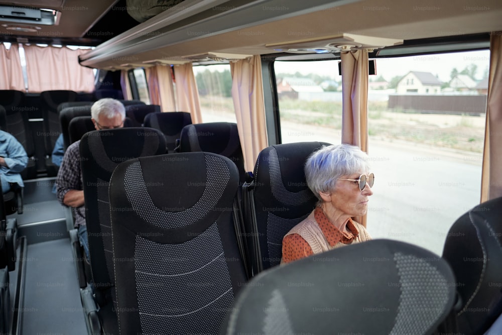 Gealterte kaukasische Frau mit grauen Haaren, die am Fenster im Bus sitzt und auf Straßen und Landhäuser schaut