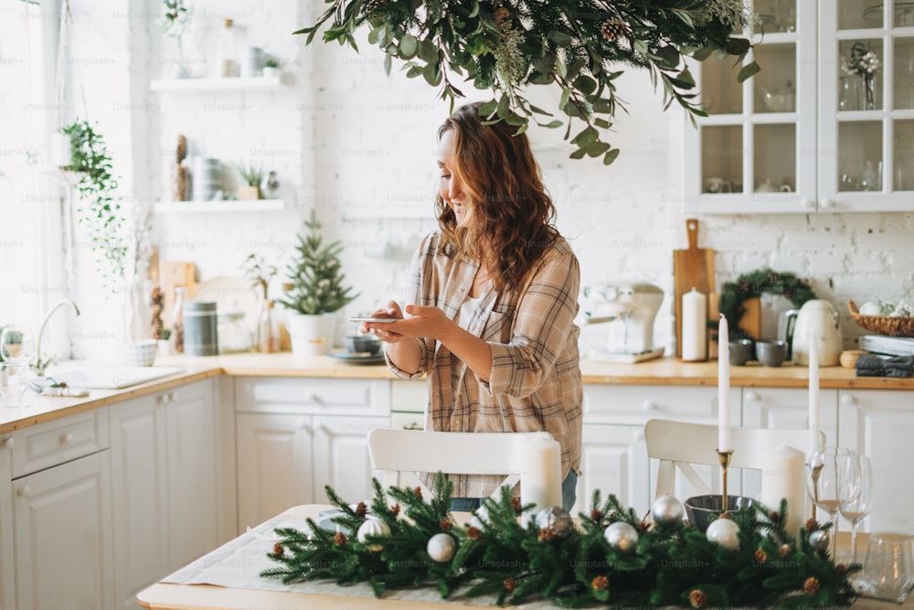 Attractive curly hair woman in plaid shirt takes photo gift box on festive table with fir composition, christmas time