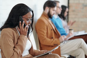 Side view of serious young Black woman sitting on chair in corridor and talking by phone while waiting for job interview