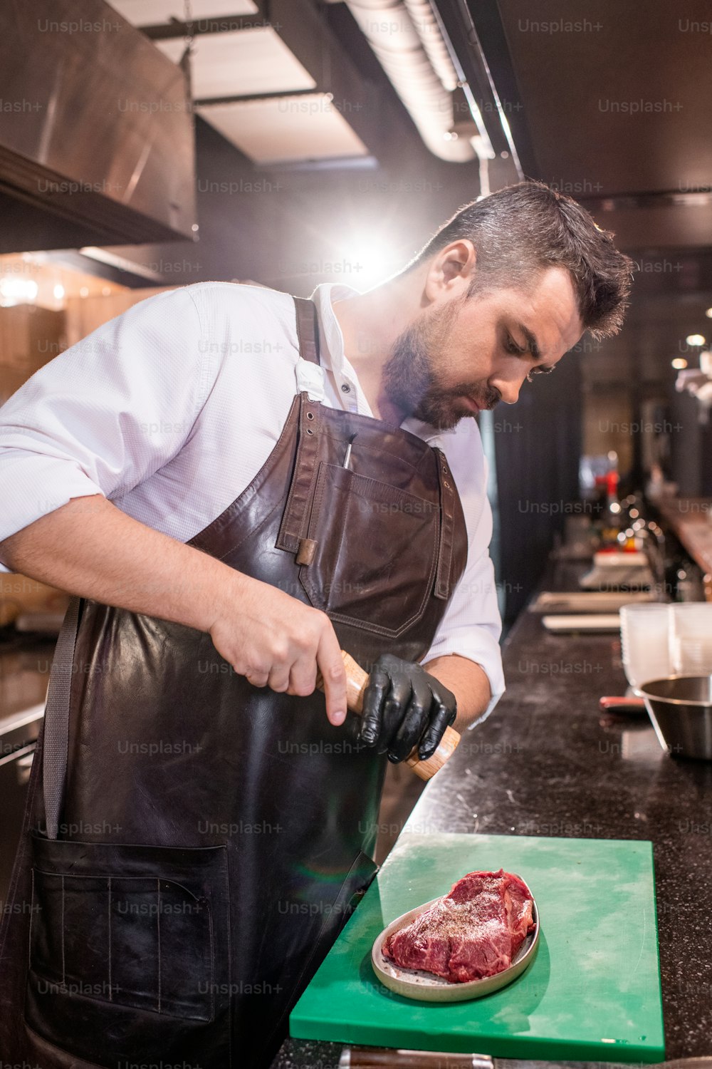 Serious bearded chef in apron peppering steak while marinating it at restaurant kitchen