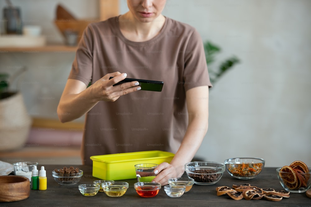 Young craftswoman with smartphone standing by table and shooting bowls containing essence oils and other stuff for making handmade soap