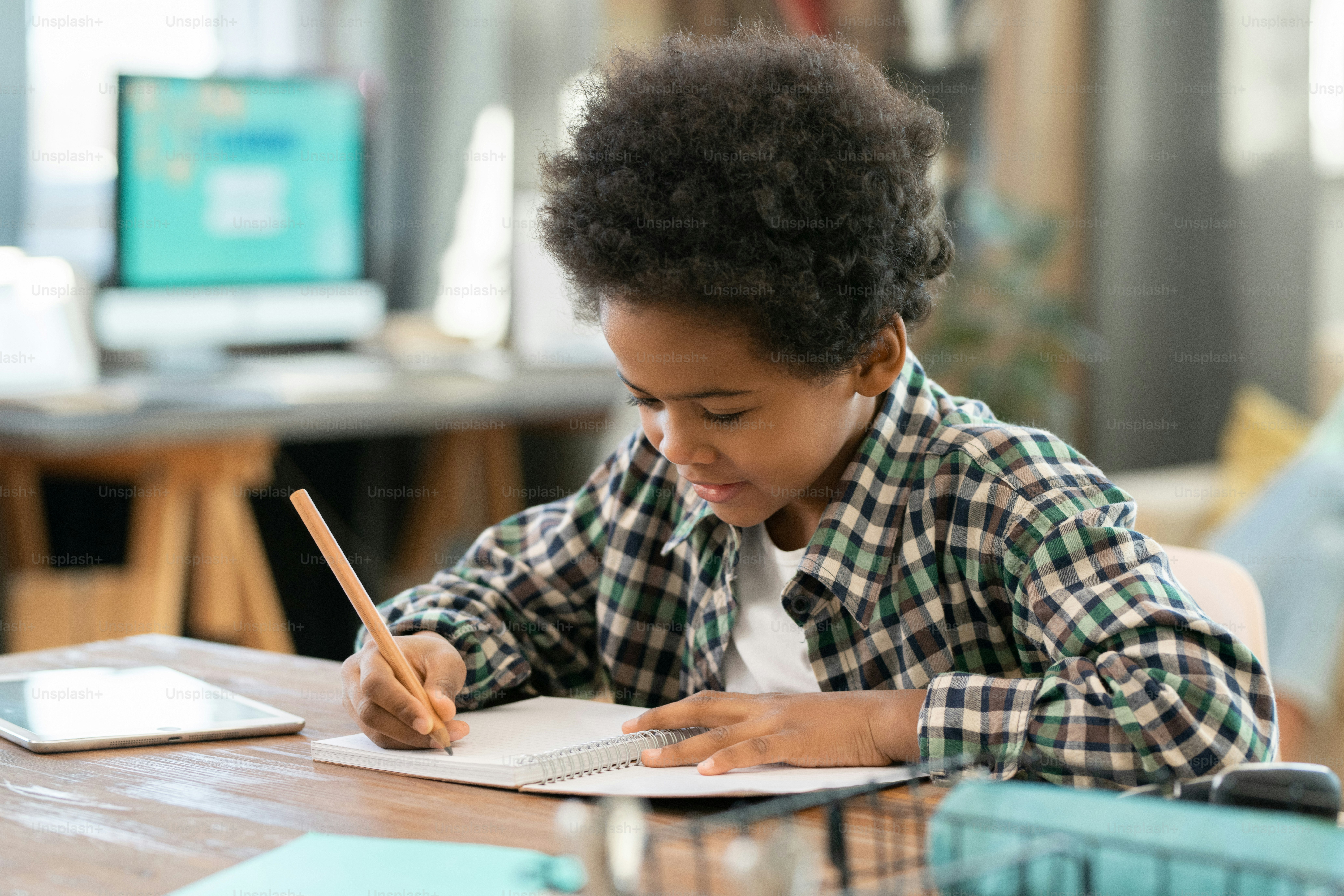 Elementary schoolboy of African ethnicity making notes in copybook while doing homework by table in living-room against computer