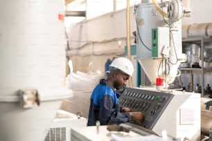 Vista lateral del joven trabajador africano con mono y casco parado frente al panel de control de una enorme máquina industrial y un mecanismo de ajuste
