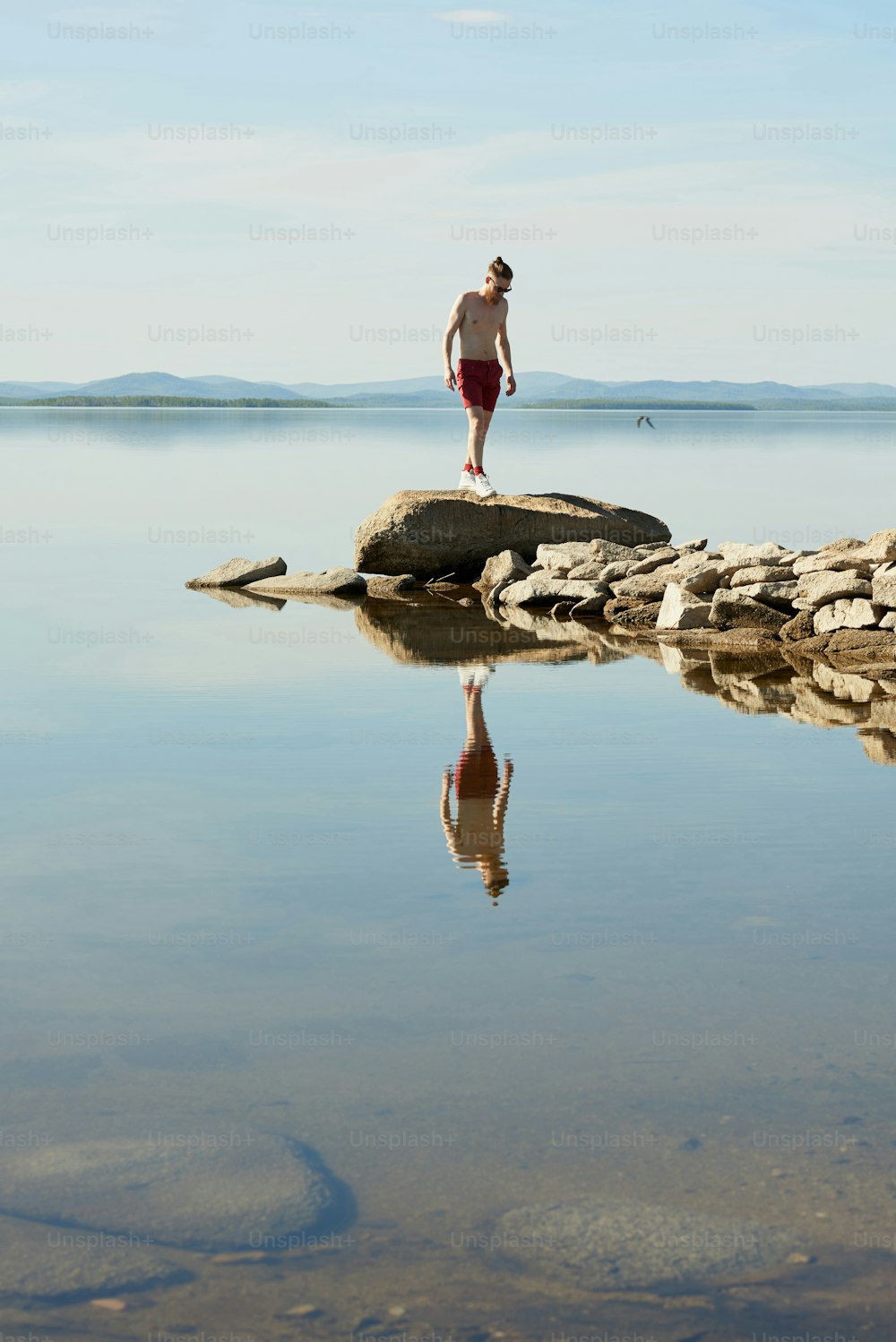 Unrecognizable young topless man walking on stones on background of picturesque calm lake and blue sky on summer afternoon